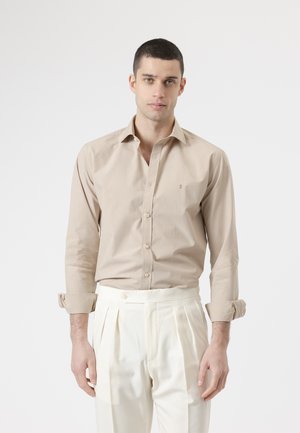 Hombre joven de frente, vistiendo una camisa beige abotonada con las mangas remangadas y pantalones blancos plisados de talle alto.