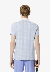 Ljusblå kortärmad polotröja med rutmönster, med en vit krage och subtil textur, visad bakifrån med en tennisrack.