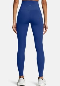Persona che indossa leggings a vita alta di colore blu reale e sneakers bianche, vista da dietro su uno sfondo neutro.