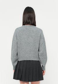 Frau mit schulterlangem, dunklem Haar, die einen grauen Strickpullover und einen schwarzen Faltenrock trägt, steht vor einem schlichten weißen Hintergrund.
