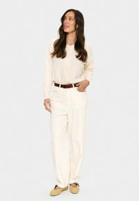 Femme aux longs cheveux noirs portant un cardigan crème boutonné, un pantalon blanc taille haute avec une ceinture marron, et des ballerines en toile beige.