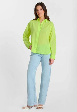 Donna in piedi che indossa una camicia verde neon con bottoni, jeans azzurri e sandali bianchi con tacco e punta aperta su uno sfondo bianco uniforme.