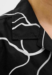 Camisa negra de manga corta con un patrón abstracto de líneas blancas, con un cuello doblado y una textura suave. Detalles de costura visibles.