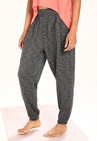 Eine Frau trägt lockere, dunkelgraue melierte Jogginghosen und ein korallenfarbenes ärmelloses Oberteil. Sie steht barfuß auf einer hellrosa Yogamatte.