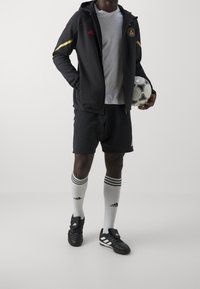 Svart sportjacka med gula och röda accenter, vit t-shirt, svarta shorts och svarta fotbollsskor. Håller en vit fotboll.