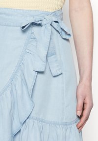 Saia envelope azul claro feita de um tecido suave, com bainha assimétrica, detalhe de laço lateral e uma silhueta fluida com seções franzidas.