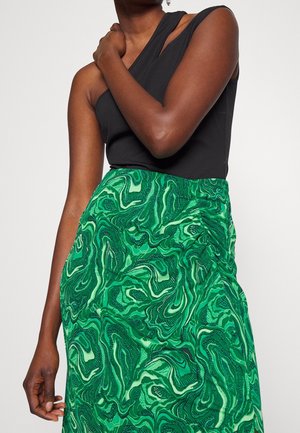 Personne portant un haut noir sans manches et une jupe verte taille haute avec un motif abstrait tourbillonnant, bras plié sur la poitrine.