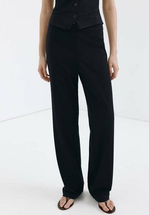 Personne portant un pantalon noir ample, un gilet noir sans manches et des tongs marron, debout sur un sol blanc uni.