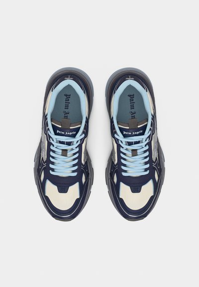 Χοντρά αθλητικά παπούτσια σε ναυτικό μπλε, κρεμ και ανοιχτό μπλε με τη σήμανση "Palm Angels" στη γλώσσα και στη σόλα, εμφανιζόμενα από πάνω σε λευκό φόντο.