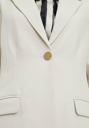 Blazer beige su misura con rever a soffietto, chiusura con un bottone caratterizzato da un design a forma di cuore e due tasche frontali. Tessuto di texture liscia.