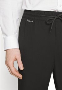 Pantalons de sport noirs avec une taille élastique et un cordon de serrage. Présente une étiquette de marque argentée sur la taille, avec une texture lisse et une coupe ajustée.