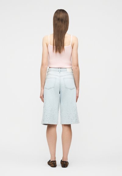 Γυναίκα που στέκεται με την πλάτη προς τα εμπρός, φοράει ροζ αμάνικη μπλούζα, γαλάζια σορτς μέχρι το γόνατο με σχέδιο πιτσιλίσματος και παπούτσια με λεοπάρ μοτίβο.