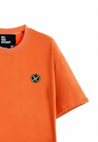 Camiseta naranja de manga corta hecha de algodón, con escote redondo y un parche circular negro con logo en el lado izquierdo del pecho.