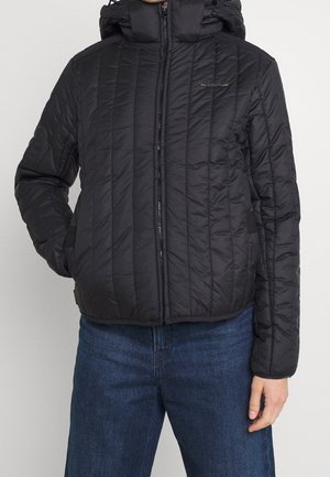 Person trägt eine schwarze gesteppte Jacke mit Reißverschluss vorne und Kapuze, kombiniert mit blauen Jeans.