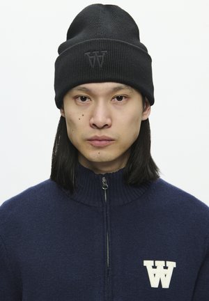Zwarte gebreide beanie met een omgeslagen boord en een reliëflogo. Model draagt een marineblauwe ritstrui met een geribde kraag en een wit geborduurd embleem.