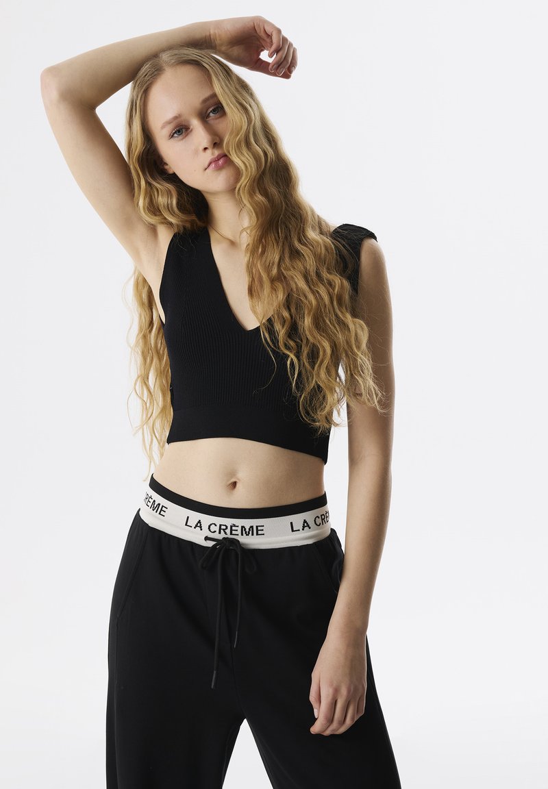 Zwarte geribbelde crop top met een diepe V-hals, gecombineerd met zwarte joggers met een witte elastische tailleband met de tekst "LA CRÈME." Lang golvend haar.