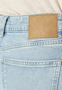 Svetlo modri jeans iz denima z zadnjim žepom iz svetlo rjave usnje, na katerem je embosirano besedilo "AWARE". Vidna so šivi in žepi.