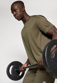 Olívazöld, rövid ujjú sportpóló fekete logóval, matt felülettel. Egy személy fekete súlytárcsát emel, jól definiált karizmokkal.