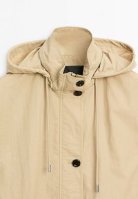 Veste à capuche beige avec col montant, boutons noirs, fermeture éclair frontale sous rabat, et cordons avec embouts métalliques sur fond blanc.