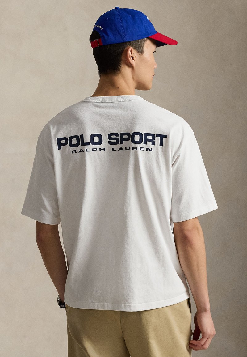 T-shirt en coton blanc avec l'inscription « POLO SPORT RALPH LAUREN » imprimée en bleu marine dans le dos. Associé à une casquette bleue et rouge, présentant un design décontracté.