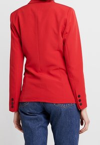 Červené sakové sako s přiléhavým střihem, jednolícím zadním rozparkem a knoflíkovými manžetami. Nosené s modrými denimovými džíny. Hladká textura, strukturovaný design.