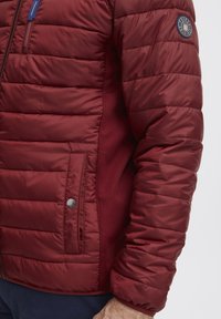 Giacca a vento bordeaux con motivo trapuntato e pannelli in rete laterali. Presenta una tasca con zip e un'etichetta con logo sulla manica. Tessuto liscio e leggero.