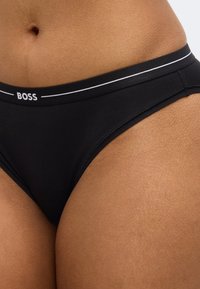 Čierne bikiny s bielym elastickým pásom s nápisom značky "BOSS." Hladká látka s priliehavým strihom, minimalistický dizajn.