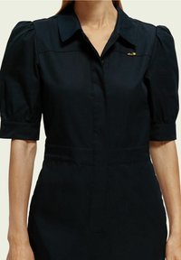 Vrouw in een zwarte jurk met opgezette halve mouwen, een kraag en een kleine gouden broche in de vorm van een planeet en ster.