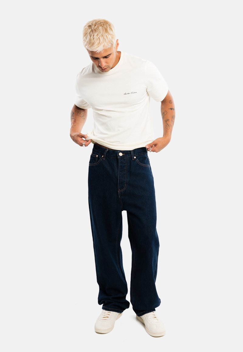 Jeune homme aux cheveux blonds courts portant un t-shirt blanc, un jean large foncé et des baskets blanches, regardant vers le bas tout en tenant l'ourlet de son t-shirt.