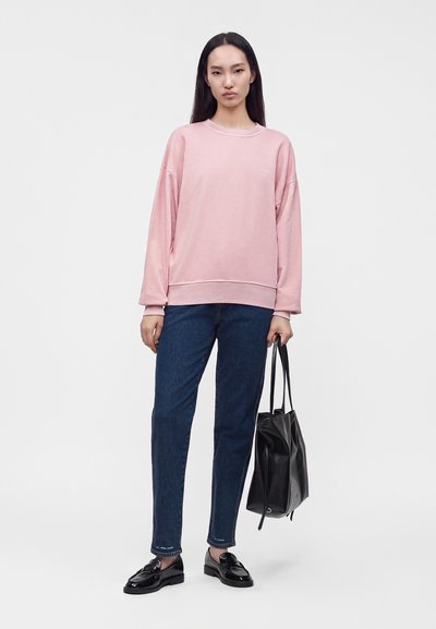 Femme aux longs cheveux foncés portant un sweat-shirt rose ample, un jean bleu, des mocassins noirs, tenant un sac cabas en cuir noir.