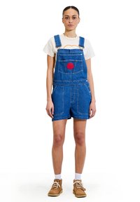 Shorts in denim blu con bretelle regolabili, tasca frontale e dettagli ricamati. Abbinati a una maglietta bianca e scarpe marroni.
