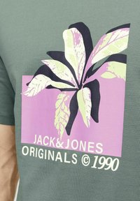 Roheline puuvillane t-särk, millel on suur graafika lehtedest kreemja, roosa ja musta värviga, ning all on tekst "JACK & JONES ORIGINALS © 1990".