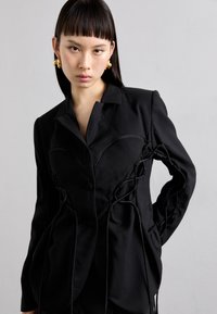 Schwarzer, maßgeschneiderter Blazer mit taillierter Passform und geschnürten Akzenten, Ein-Knopf-Verschluss und dezenten, geschwungenen Nähten auf der Vorderseite.