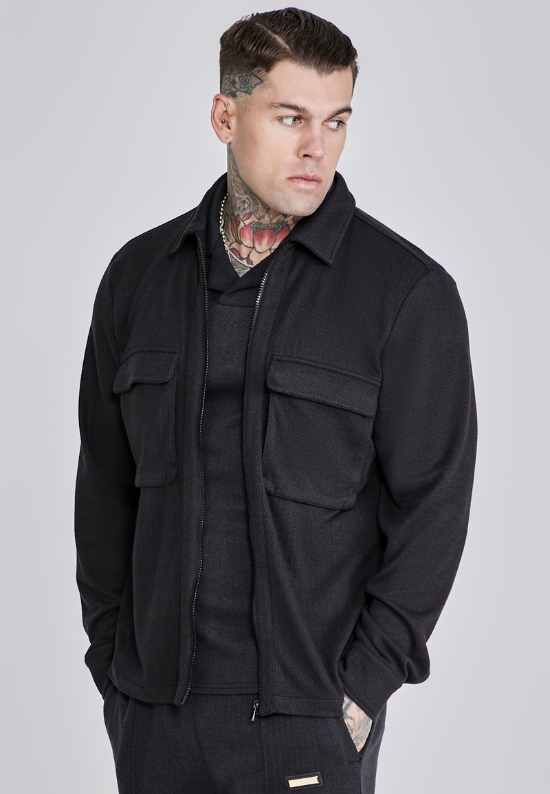 Chaqueta negra con cremallera y tejido texturizado, con dos bolsillos frontales y un cuello. Lleva puesta sobre una camiseta negra de manga corta, con un acabado suave.