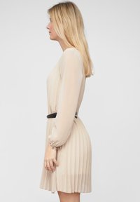 Robe plissée beige à manches longues, avec une ceinture noire à la taille et une texture douce et transparente. La vue de côté montre un drapé délicat.