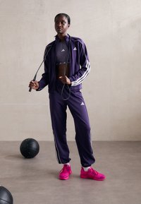 Lilla treningsdrakt med hvite striper, kombinert med en grå crop top. Lyse rosa joggesko. Modell som holder et hoppetau.