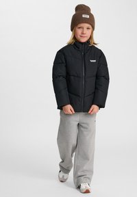 Svart puffjacka med dragkedja framtill och sömdetaljer, som kombineras med grå sweatpants och en brun stickad beanie. Vita sneakers med detaljer.