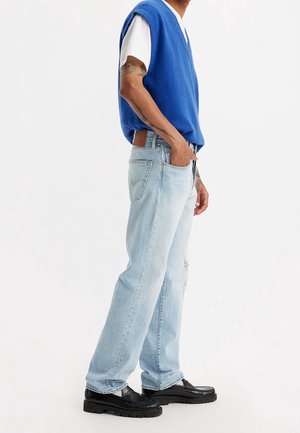 Homme portant un pull sans manches bleu par-dessus un T-shirt blanc, un jean bleu clair, des mocassins noirs et des tatouages visibles sur les bras, debout devant un fond blanc.