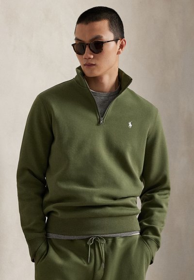 Ελιά πράσινο φούτερ με φερμουάρ στον γιακά, ριμπ λεπτομέρειες και μικρό κεντητό λογότυπο στο στήθος. Απαλή υφή υφάσματος.