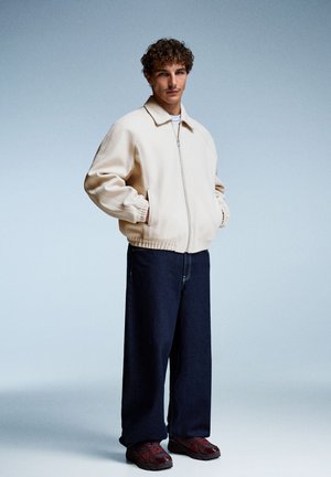 Jachetă bomber bej cu fermoar și detalii cu nervuri, combinată cu blugi închis la culoare, lejeri, și adidași vișinii. Modelul stă cu mâinile în buzunare.