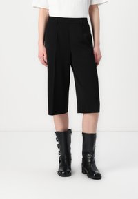 Pantaloni negri scurți cu o croială ajustată, având pliuri și o textură netedă, asortați cu cizme negre din piele până la genunchi, cu catarame.