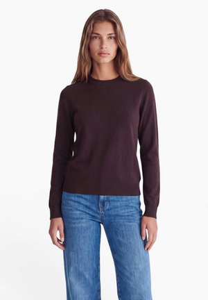 Junge Frau mit langen Haaren, die einen dunkelbraunen Pullover mit langen Ärmeln und blaue Jeans trägt, steht vor einem weißen Hintergrund.
