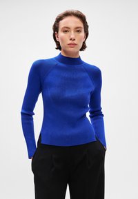 Sweater de malha canelada em azul vivo com gola alta, mangas longas e aberturas laterais; o design justo realça a silhueta.