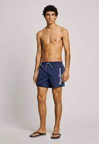 Ung man som står utan skjorta iförd marinblå badshorts med vit "Pepe Jeans"-logotyp och svarta flip-flops framför en enfärgad bakgrund.