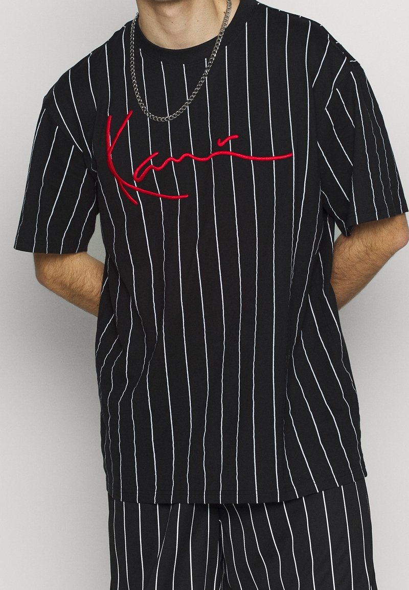 Czarny t-shirt w paski z bawełny, z białymi pionowymi paskami, z czerwonym haftowanym logo na wysokości klatki piersiowej. Krótkie rękawy, luźny krój.