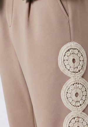 Beige broek met zijzakken, voorzien van drie witte, cirkelvormige gehaakte kantapplicaties die verticaal naast de dij zijn uitgelijnd.