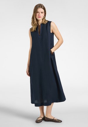 Kvinde med skulderlangt hår iført en navy ærmeløs midi-kjole med sidelommer og sorte flettede flade sko, stående mod en ensfarvet baggrund.