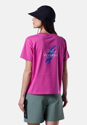 Donna che indossa una t-shirt rosa con la scritta "Trail Memories", pantaloncini verdi con tasca nera e un cappello a tesa larga nero, vista da dietro su uno sfondo semplice.