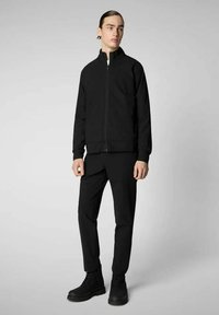 Giacca nera con zip e collo alto, abbinata a pantaloni coordinati e stivali neri con lacci. Texture liscia; design minimalista.