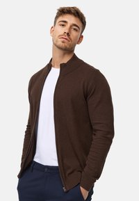 Braune Zip-Kardigan mit geripptem Kragen, langen Ärmeln und tailliertem Design, getragen über einem weißen T-Shirt und kombiniert mit blauen Hosen.